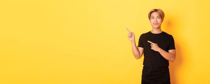 Portrait of smiling handsome asian man in black t-shirt pointing fingers upper left corner, showing logo or banner, yellow background.