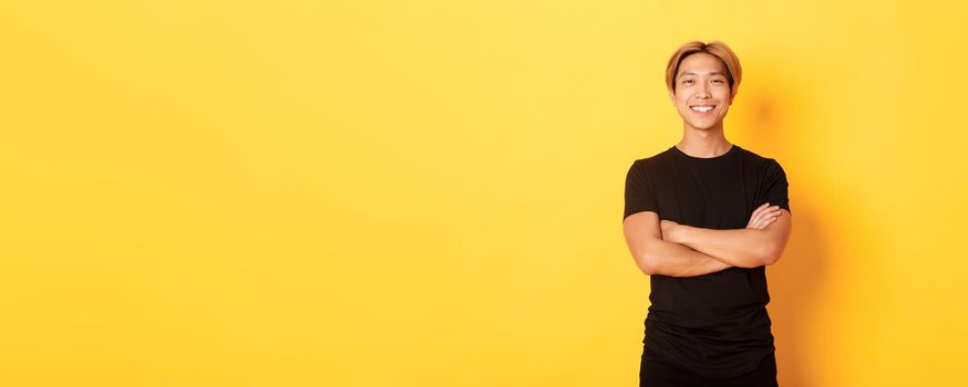 Portrait of confident handsome asian man smiling satisfied, standing over yellow background in black clothes.