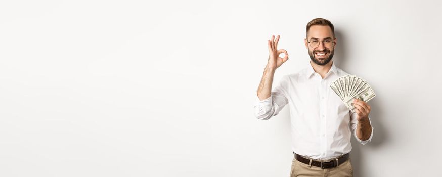 Satisfied young businessman showing money, make okay sign, earning cash, standing over white background.