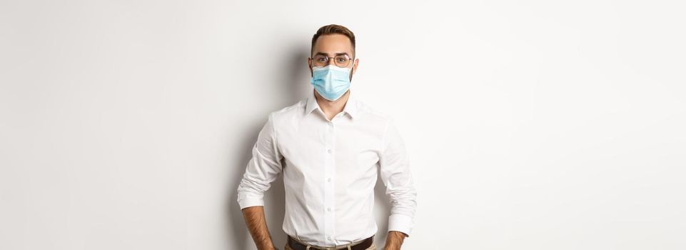 Covid-19, social distancing and quarantine concept. Male employee wearing face mask for work, standing against white background.