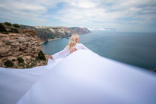 Blonde with long hair on a sunny seashore in a white flowing dress, rear view, silk fabric waving in the wind. Against the backdrop of the blue sky and mountains on the seashore