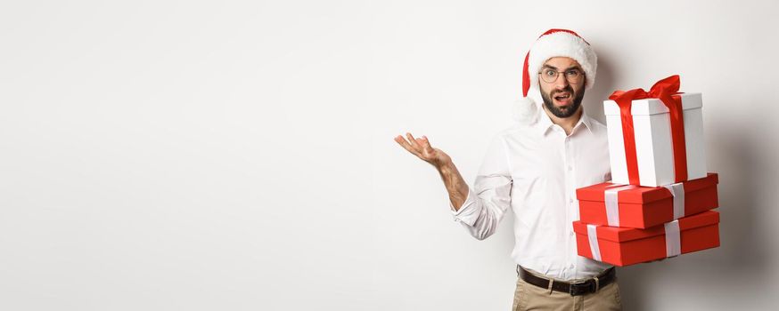 Merry christmas, holidays concept. Man looking confused while holding xmas gifts, shrugging puzzled, standing in santa hat against white background.