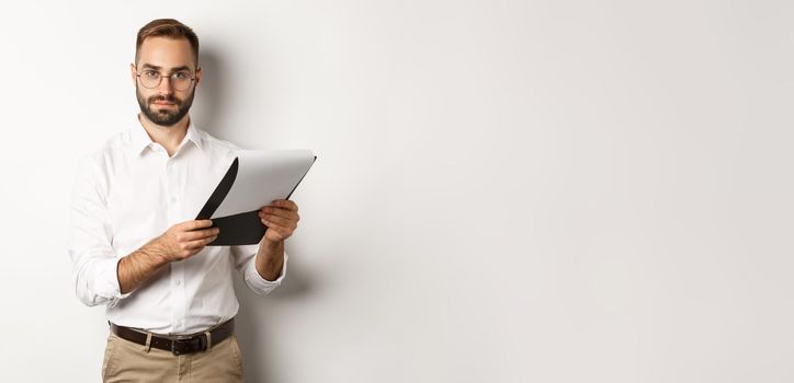 Serious employer looking at camera while reading documents on clipboard, having job interview, standing over white background.