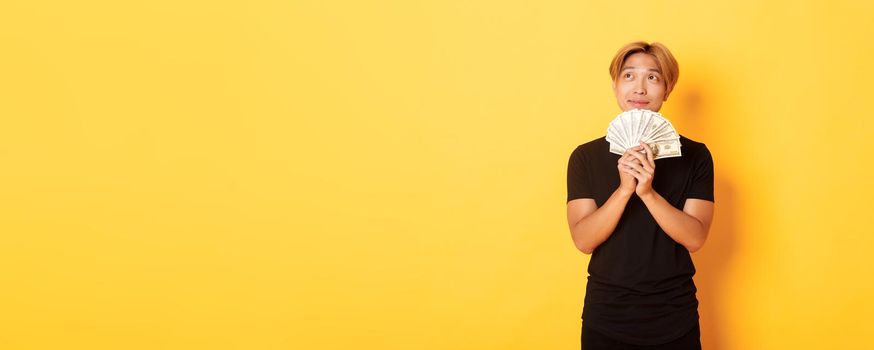 Portrait of dreamy handsome asian guy showing his savings and thinking, looking upper left corner, holding money, standing yellow background.