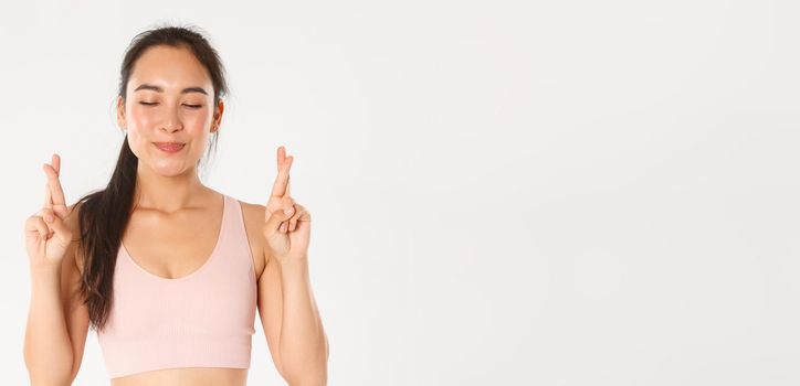 Sport, wellbeing and active lifestyle concept. Close-up of smiling optimistic asian girl, hope lose weight, cross fingers for good luck and close eyes while making wish, white background.