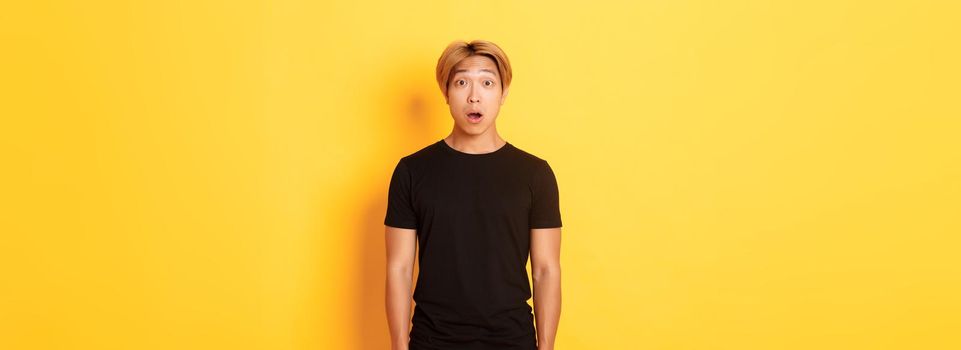 Portrait of startled handsome asian blond guy, drop jaw and gasping impressed, yellow background.