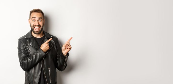 Excited, handsome man in black leather jacket, pointing fingers right at promo offer, showing logo, white background.