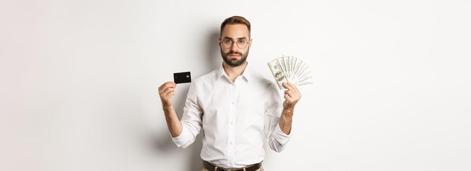 Serious businessman looking at camera, holding credit card and money, standing over white background. Concept of shopping and finance.