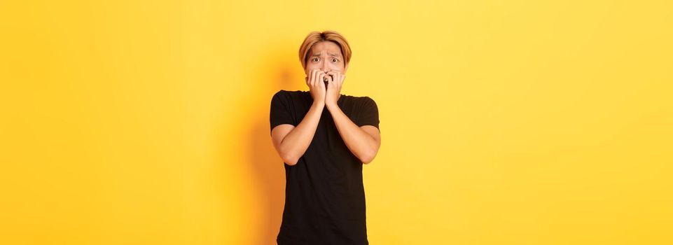 Portrait of scared insecure asian blond guy, holding hands over mouth horrified, looking frightened, yellow background.