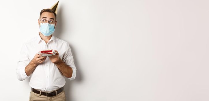 Covid-19, social distancing and celebration. Thoughtful man holding birthday cake, making wish and wearing face mask on quarantine, white background.