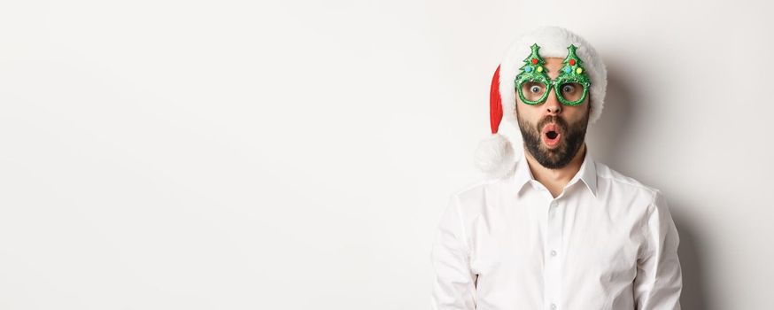 Adult man celebrating winter holidays, wearing christmas party glasses and santa hat, looking surprised at camera, standing over white background.