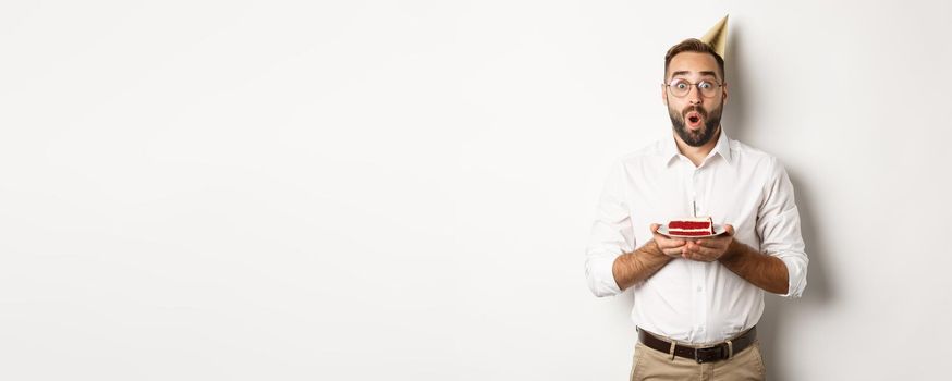 Holidays and celebration. Happy man having birthday party, making wish on b-day cake and smiling, standing against white background.