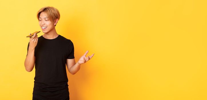 Portrait of stylish handsome korean guy with blond hair record voice message on mobile phone, holding smartphone near mouth and speaking, standing yellow background.