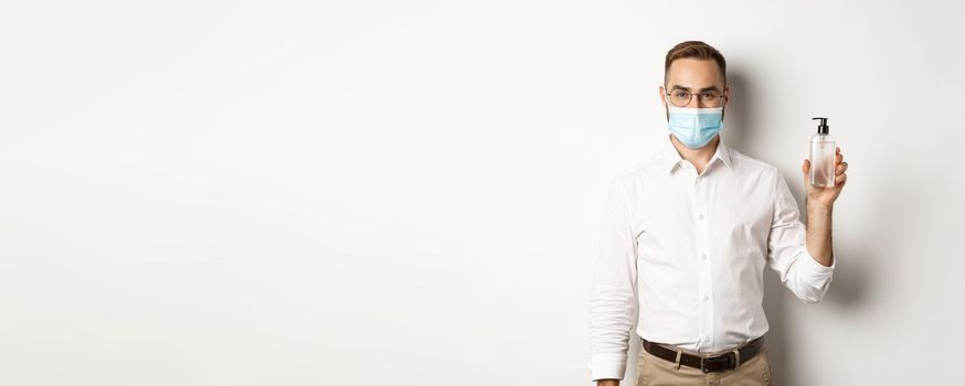Covid-19, social distancing and quarantine concept. Employer in medical mask showing hand sanitizer, asking to use antiseptic at work, standing over white background.