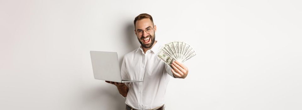 Business and e-commerce. Happy successful businessman bragging with money, working on laptop online, standing over white background.