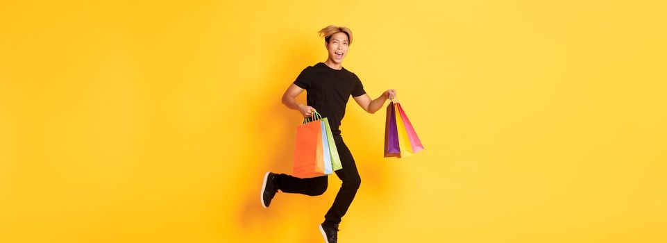 Full-length of happy attractive asian man jumping from happiness and carry shopping bags, standing yellow background.