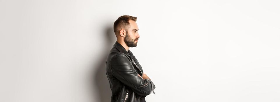 Profile of handsome serious man in leather jacket, looking left, holding hands on chest confident, white background.