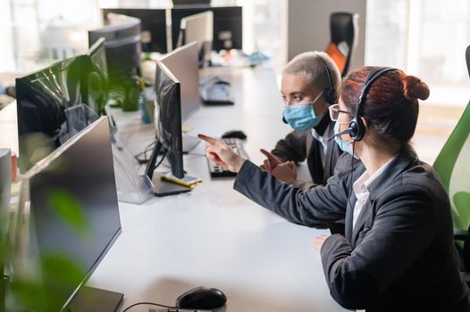 Two women in medical masks and headsets are working in the office.