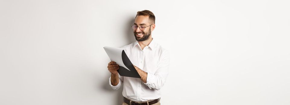 Man looking satisfied while reading documents, holding clipboard and smiling, standing over white background.