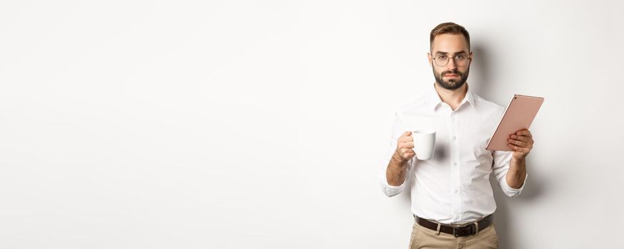 Confident male manager reading work on digital tablet and drinking coffee, standing over white background.