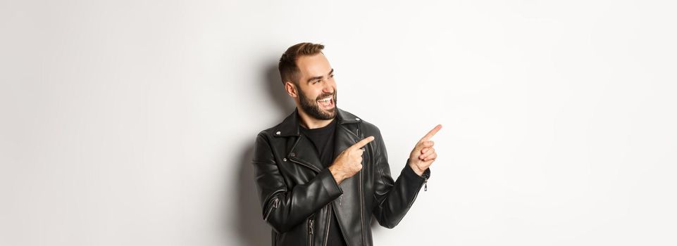 Confident macho man in black leather jacket, pointing fingers left at promo offer, showing logo, white background.