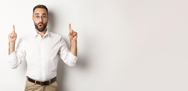 Excited handsome guy in glasses pointing fingers up, showing information banner, standing over white background. Copy space