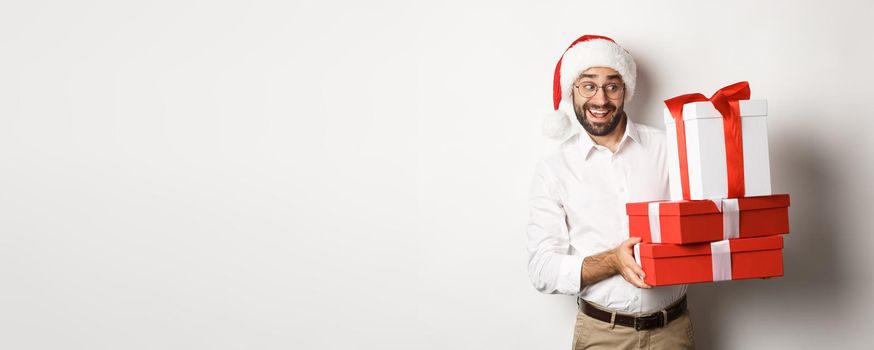 Merry christmas, holidays concept. Excited man celebrating xmas, wearing santa hat and holding gifts, standing over white background.