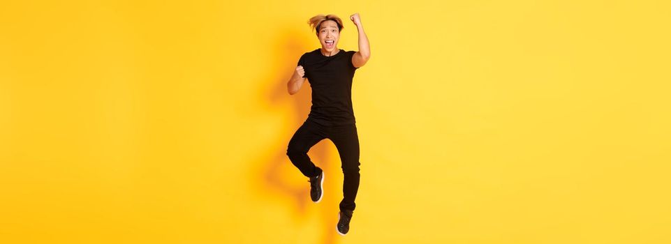 Full-length of happy attractive asian guy in black clothes jumping and celebrating victory, achieve goal, standing yellow background.