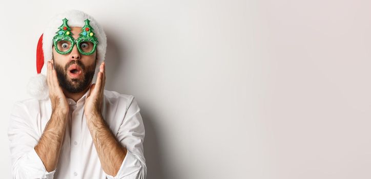 Adult man celebrating winter holidays, wearing christmas party glasses and santa hat, looking surprised at camera, standing over white background.