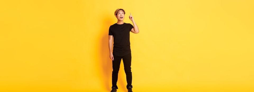 Full-length of thoughtful handsome asian man, raising finger up, have good idea, standing yellow background.