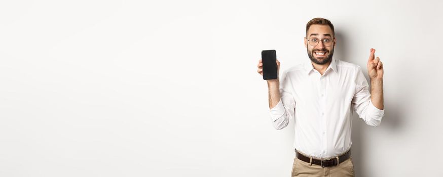 Hopeful young business man showing mobile screen, holding fingers crossed, waiting for online results, standing over white background.