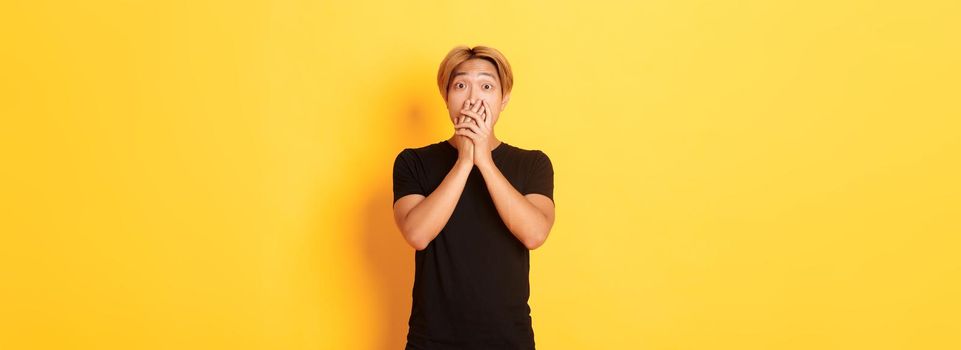 Portrait of surprised asian handsome guy with blond hair, cover mouth with hands and stare at camera impressed, yellow background.