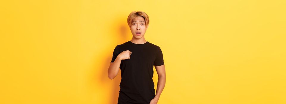 Portrait of confused and sad asian blond guy, frowning upset and pointing at himself, standing yellow background.