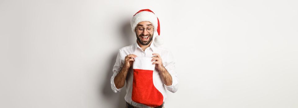 Happy adult man open christmas sock and looking inside, receiving xmas gifts for winter holidays, standing in santa hat, white background.