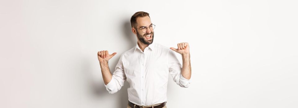 Satisfied and self-assured businessman pointing at himself, standing over white background.