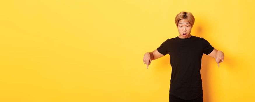 Portrait of surpirsed asian guy gasping amazed, pointing fingers and looking down at banner, standing yellow background.