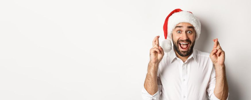 Party, winter holidays and celebration concept. Happy man in santa hat making christmas wish, cross fingers for good luck and looking excited, white background.