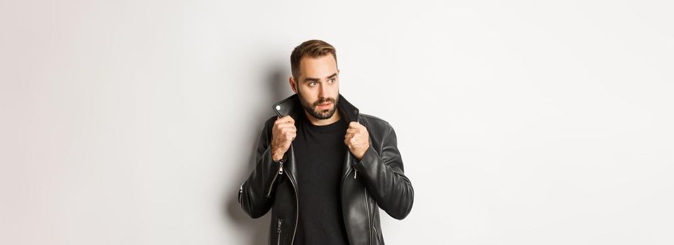 Attractive bearded man in leather biker jacket looking aside, standing confident against white background.