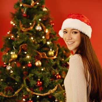 Let the festivities begin. A beautiful young woman wearing a santa hat standing by a Christmas tree