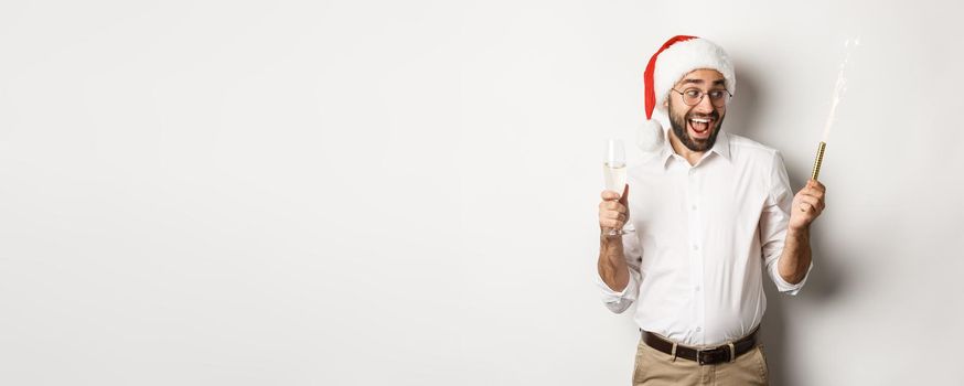 Winter holidays and celebration. Excited man celebrating New Year eve with fireworks sparkles and drinking champagne, wearing santa hat, white background.