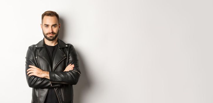 Attractive macho man in leather jacket smiling, looking confident with hands crossed on chest, white background.