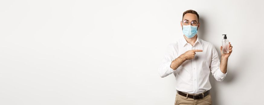 Covid-19, social distancing and quarantine concept. Office worker in medical mask pointing at hand sanitizer, showing antiseptic, white background.