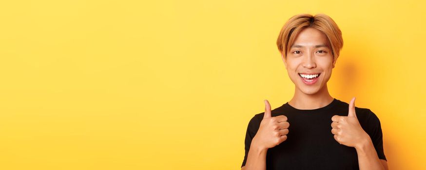 Close-up of satisfied, happy asian guy in black t-shirt, showing thumbs-up in approval, smiling and agree, standing yellow background.