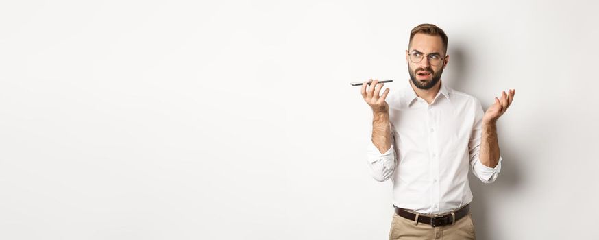 Man recording voice message or talking on speakerphone, looking confused, standing over white background.