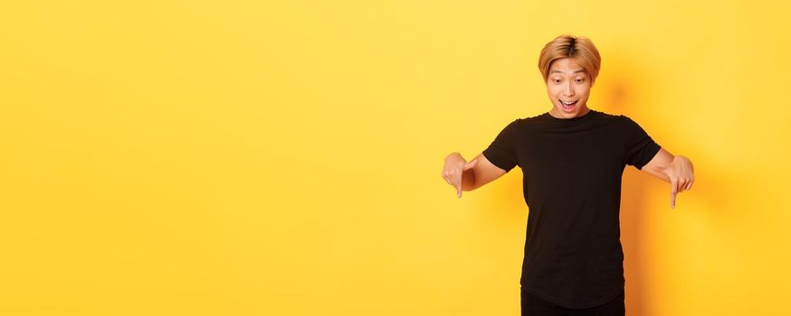 Portrait of excited and happy asian guy with blond haircut, pointing and looking down amused, standing yellow background.