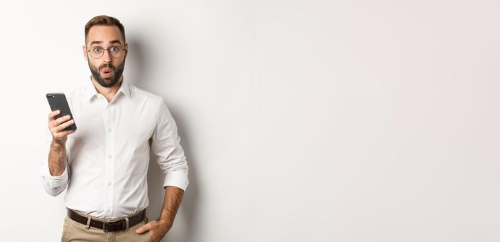 Surprised handsome man reading interesting info online, holding smartphone, standing over white background.