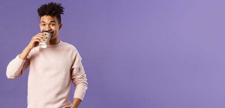 Leisure, lifestyle and people concept. Portrait of young cheerful guy drinking coffee take-away from favorite cafe, smiling enjoying nice spring day, standing purple background.