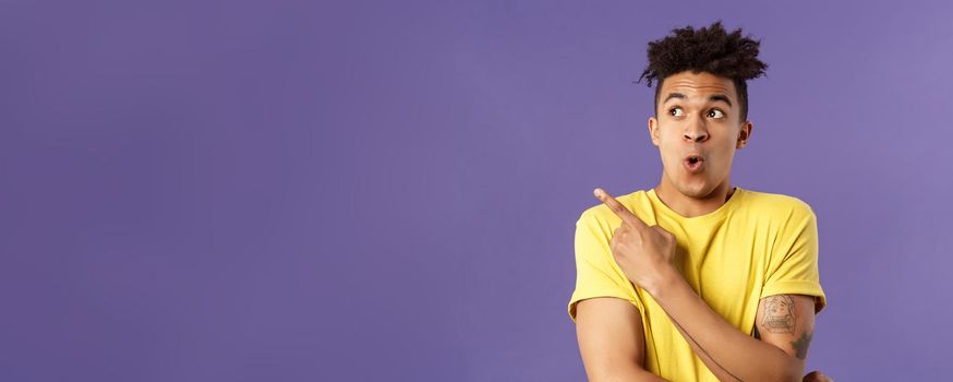 Waist-up portrait of amazed, impressed hispanic male with dreads, seeing something incredible and cool, pointing finger looking upper left corner, breathtaking awesome thing, purple background.