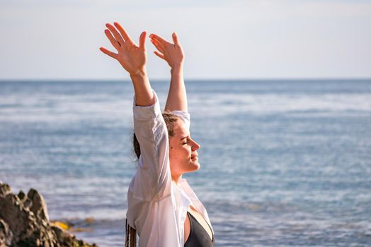 The girl stands on the shore and looks at the sea. Her hands are raised up. She wears a white shirt and her hair is in a braid