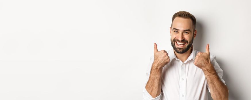 Close-up of satisfied bearded guy in white shirt, showing thumbs up in approval, like and agree, positive answer, white background.